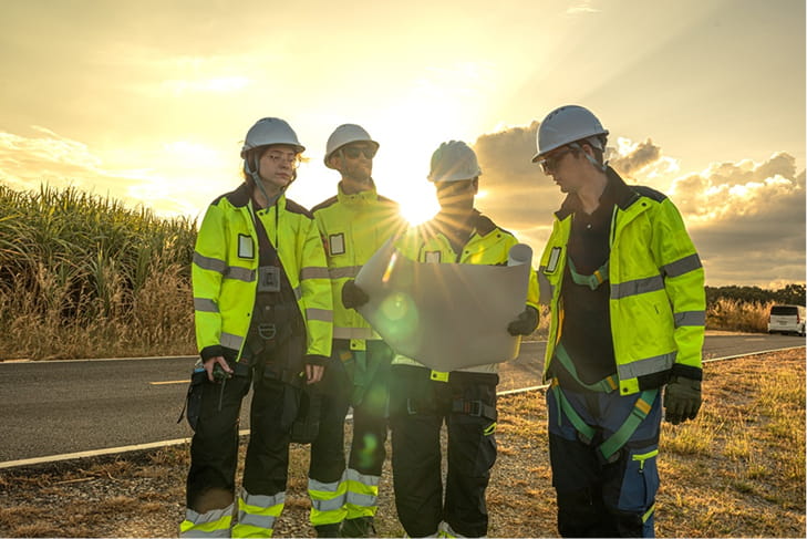 Four construction workers in safety gear, including hard hats and reflective jackets, are gathered around a blueprint in front of a sunset.